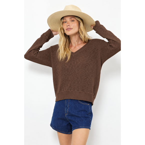 A woman wearing a brown long-sleeve V-neck knit sweater, blue denim shorts, and a beige wide-brim hat, posing against a white studio background.