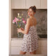 A woman wearing a white spaghetti-strap nightgown with black polka dots stands in a kitchen holding a vase of pink lilies.