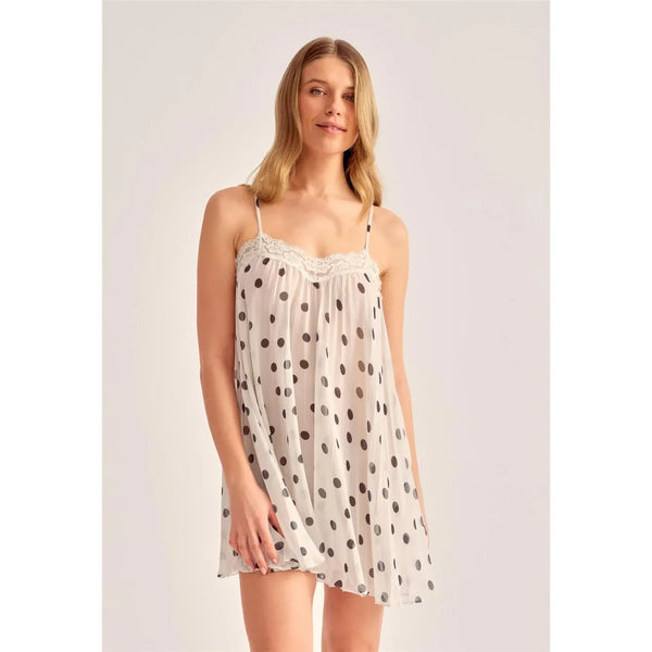 A woman modeling a white semi-sheer slip dress with black polka dots and a lace-trimmed neckline, photographed against a plain light gray studio background.