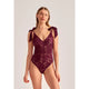 A woman modeling a deep burgundy lace bodysuit with shoulder tie details, photographed against a solid light gray studio background.