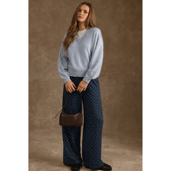 A woman stands against a textured brown studio backdrop wearing a light blue fuzzy V-neck sweater, navy wide-leg pants with small white dots, black shoes, and carrying a small brown shoulder bag.