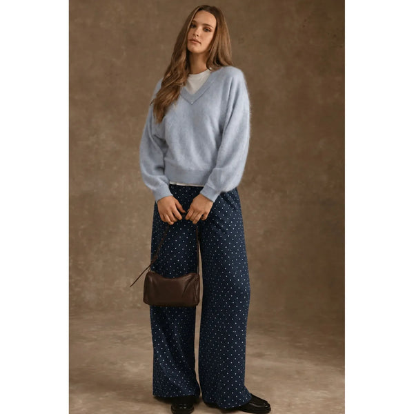 A woman stands against a textured brown studio backdrop wearing a light blue fuzzy V-neck sweater, navy wide-leg pants with small white dots, black shoes, and carrying a small brown shoulder bag.