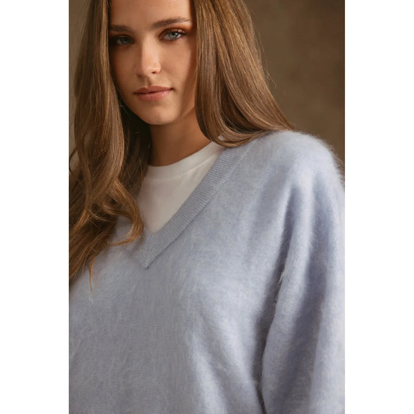 A close-up of a woman wearing a soft, light blue-gray fuzzy V-neck sweater over a white crewneck top, set against a warm brown background.