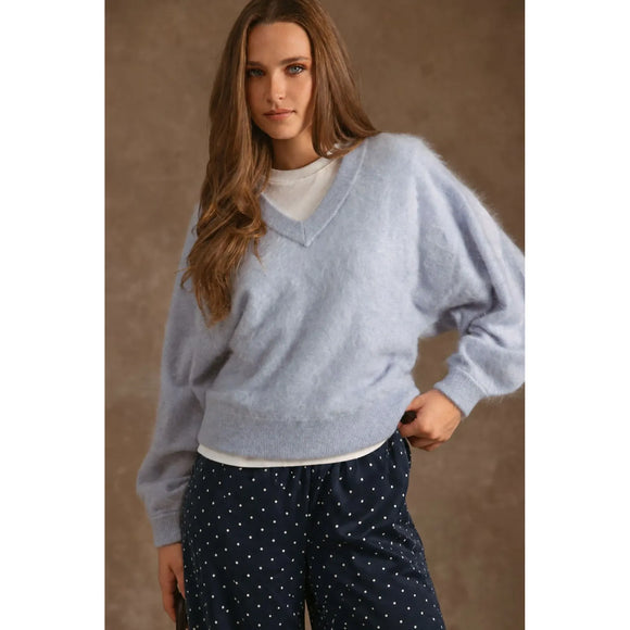 A woman wearing a light blue fuzzy V-neck sweater over a white top and navy polka-dot pants, standing against a textured brown studio backdrop.