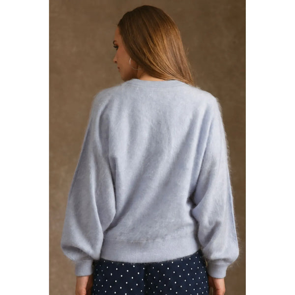 A woman viewed from the back wearing a light blue-gray fuzzy knit sweater and navy polka-dot pants against a textured brown studio backdrop.