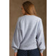 A woman viewed from the back wearing a light blue-gray fuzzy knit sweater and navy polka-dot pants against a textured brown studio backdrop.