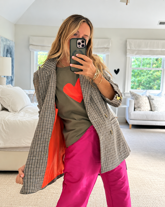 A woman takes a mirror selfie in a bright bedroom, wearing an oversized black-and-white houndstooth blazer with a bright orange lining, a green graphic t-shirt with a red heart, and hot pink pants.