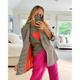 A woman takes a mirror selfie in a bright bedroom, wearing an oversized black-and-white houndstooth blazer with a bright orange lining, a green graphic t-shirt with a red heart, and hot pink pants.