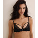 A woman modeling a black lace underwire bra with thin adjustable shoulder straps and small gold-tone ring details at the shoulder attachments, photographed against a plain light beige wall.