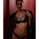 A model wearing a black lace bra and matching panties, with a black satin jacket draped open over her shoulders, posed against a dark red textured wall.