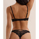 Rear view of a model wearing a black lace bralette and matching lace thong with gold-tone hardware against a warm beige studio background.
