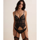 A woman modeling a black lace lingerie set with a sheer corset-style top, matching panties, and attached thigh straps against a plain light beige studio background.
