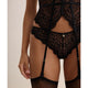 Close-up of a model wearing a black lace garter belt and matching lace panties with attached suspenders and sheer black thigh-high stockings against a plain beige background.