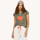 A woman modeling an olive green short-sleeve t-shirt with a large orange-red heart on the front, paired with white sweatpants and a gray knit beanie, against a solid off-white background.