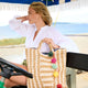 Woman sitting in a golf cart with a straw bag featuring colorful pom-poms, beach setting.