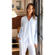 A woman wearing a light blue long-sleeve button-front pajama shirt and matching light blue pajama pants, standing indoors near a window.