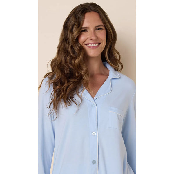 A woman with long wavy brown hair wearing a light blue pajama shirt with white piping, shown against a solid beige background.