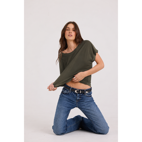 A woman kneeling on a light gray studio floor wearing an olive green oversized short-sleeve crop top and blue jeans with a black belt.