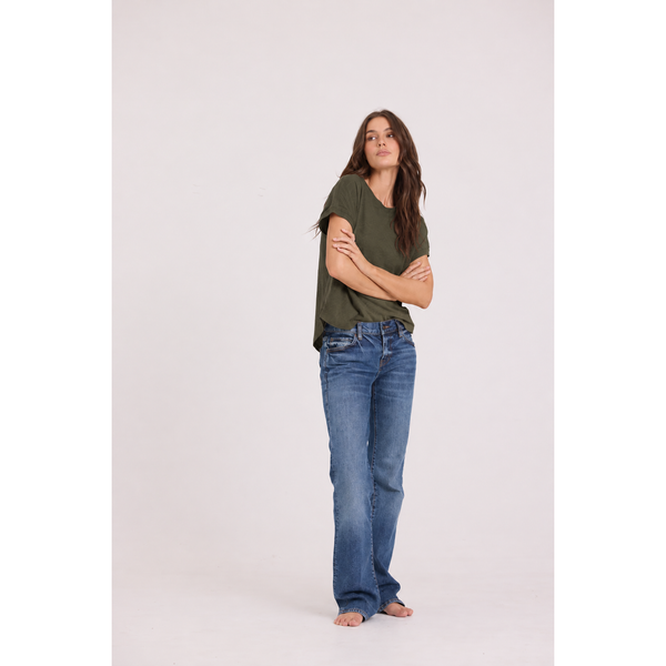 A woman stands barefoot against a plain light gray studio background, wearing a loose olive green short-sleeve top and blue jeans with a slight flare at the hem.
