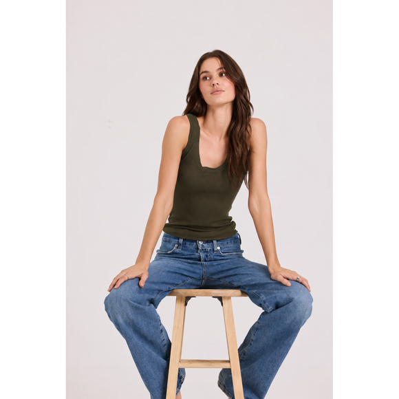 A woman sits on a wooden stool wearing an olive green sleeveless tank top and blue jeans against a plain light gray studio background.