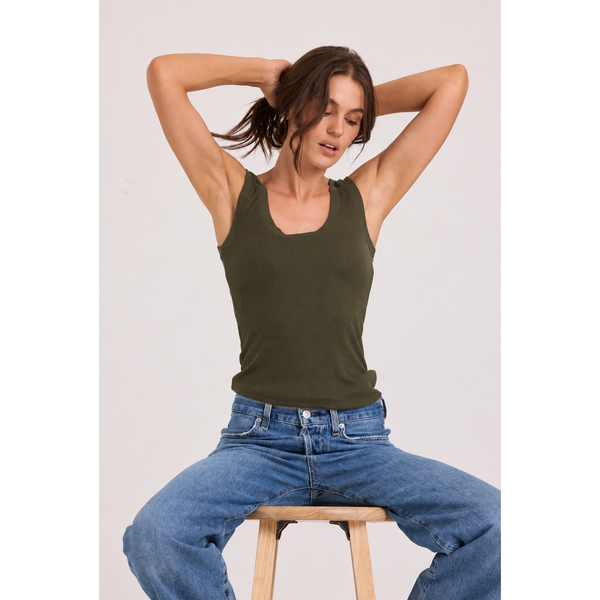 A woman wearing an olive green tank top and blue jeans sits on a wooden stool against a light gray studio background.