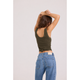 A woman shown from behind wearing an olive green sleeveless crop top and blue jeans, with her arms raised over her head, against a plain light gray studio background.
