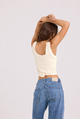 A woman shown from behind wearing a cream-colored ribbed tank top tied at the waist and blue jeans, posing with her arms raised against a plain light gray background.