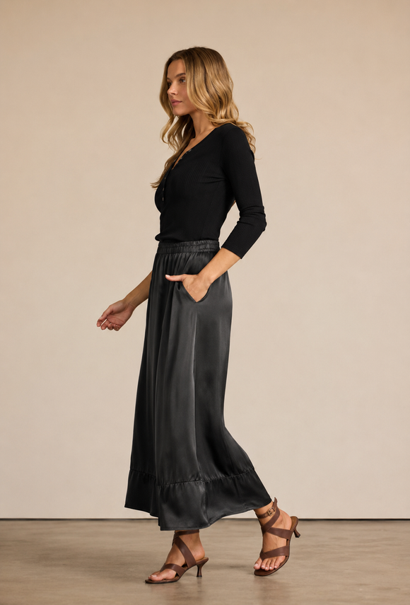 A woman modeling a black long-sleeve top and a high-waisted, flowing black midi skirt with pockets, paired with brown strappy heels, against a solid beige studio background.