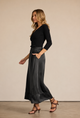 A woman modeling a black long-sleeve top and a high-waisted, flowing black midi skirt with pockets, paired with brown strappy heels, against a solid beige studio background.