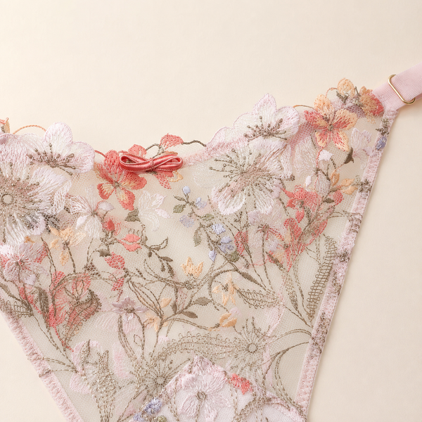 A close-up of a lace lingerie bottom with floral embroidery in soft pink, peach, white, and muted blue on a pale blush background, with a small pink bow at the center and a strap visible at the upper right.