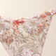 A close-up of a lace lingerie bottom with floral embroidery in soft pink, peach, white, and muted blue on a pale blush background, with a small pink bow at the center and a strap visible at the upper right.