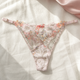 A pair of light pink lace thong panties with floral embroidery and a small bow at the center, laid flat on a textured cream linen fabric with soft pink satin fabric in the corner.