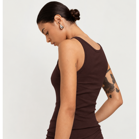 A woman wearing a dark brown ribbed tank top, shown from a side-back angle against a plain light gray studio background.