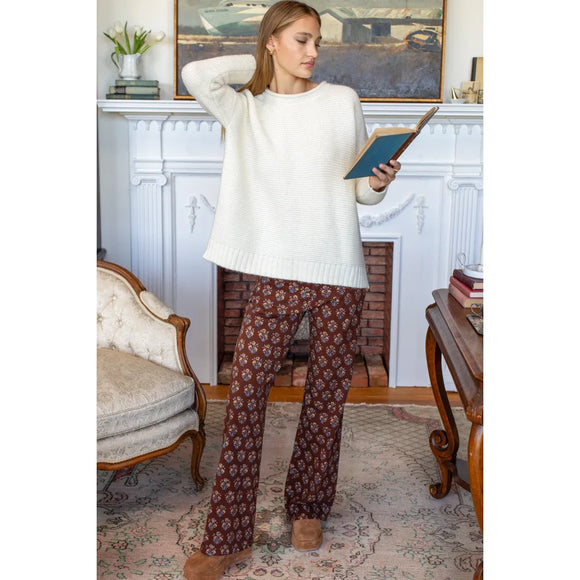 A woman stands in a living room in front of a white fireplace, wearing a cream ribbed knit sweater and brown patterned flared pants, holding an open book.