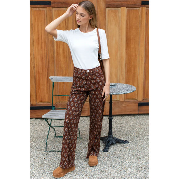 A woman stands outdoors in front of a wooden door wearing brown patterned pants with a white t-shirt and tan boots, carrying a brown shoulder bag.