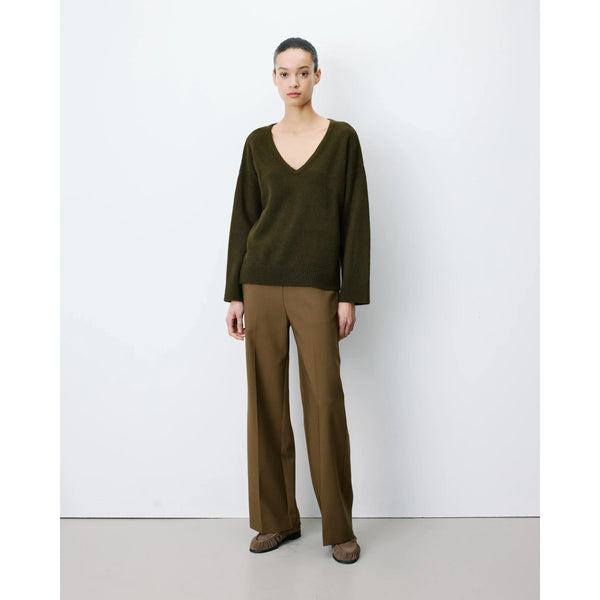 A model stands against a plain white studio wall wearing an olive green long-sleeve V-neck sweater, brown wide-leg trousers, and brown shoes.