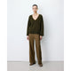 A model stands against a plain white studio wall wearing an olive green long-sleeve V-neck sweater, brown wide-leg trousers, and brown shoes.