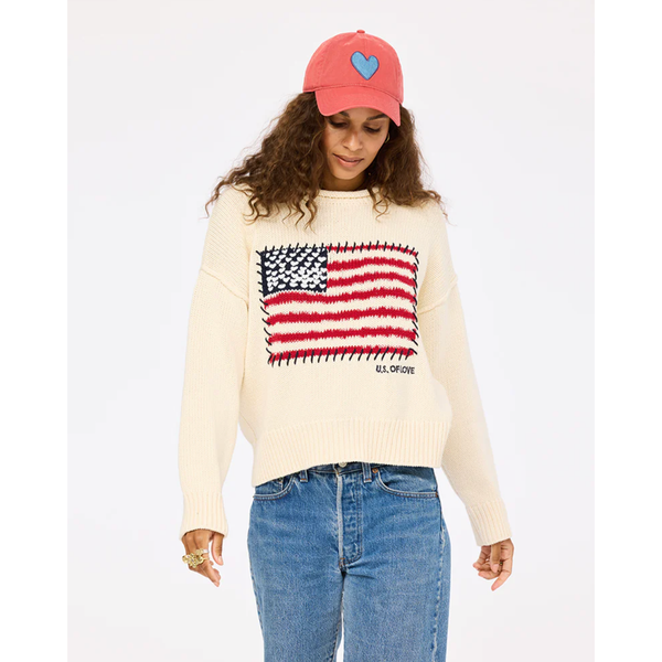 Person wearing a cream sweater with an American flag design, blue jeans, and a pink cap with a heart on a white background.