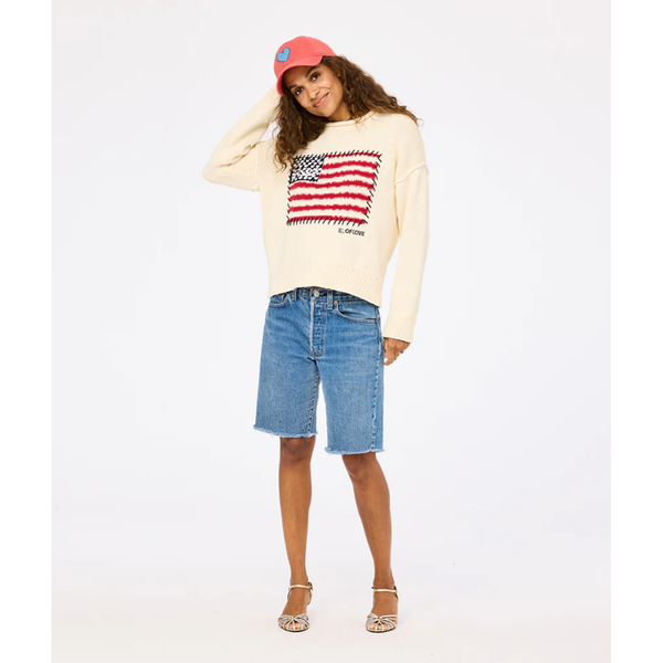 Person wearing a cream sweater with an American flag design, denim shorts, and a red cap on a white background