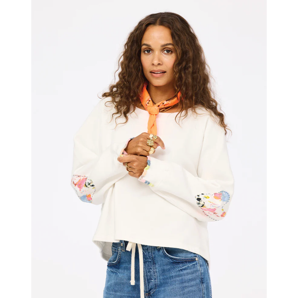 Woman wearing a white sweater with colorful sleeves and blue jeans, holding an orange scarf.