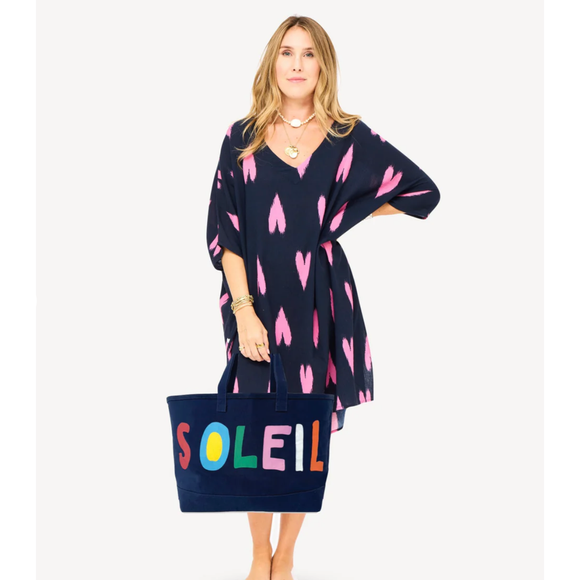Woman wearing a navy dress with pink heart patterns, holding a navy tote bag with 'SOLEIL' in colorful letters.