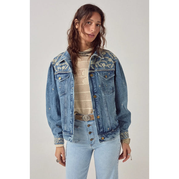 Woman wearing a blue denim jacket with floral embroidery on a plain background