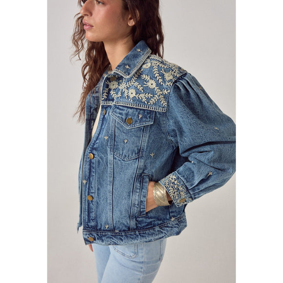 Woman wearing a blue denim jacket with floral embroidery on a plain background