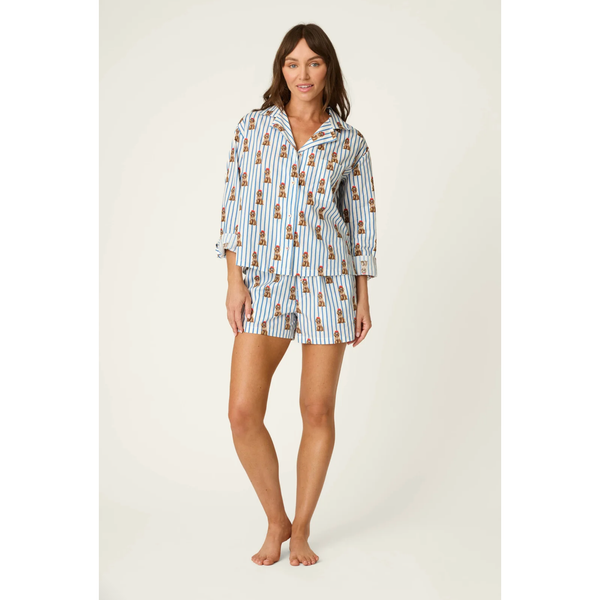 A woman modeling a matching two-piece pajama set with a white base, blue vertical stripes, and a repeating teddy bear print, photographed against a plain light beige studio background.