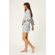 A woman modeling a matching two-piece pajama set consisting of a long-sleeve button-up shirt and shorts, featuring a blue and white striped pattern with small teddy bear prints, against a plain off-white studio background.