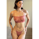 A woman modeling a dusty rose pink lace lingerie set consisting of a sheer bralette with scalloped edges and matching thong, standing against a neutral beige paneled wall.