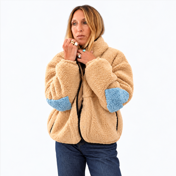 A woman wearing a beige sherpa fleece jacket with light blue elbow patches, paired with blue jeans, photographed against a solid white background.