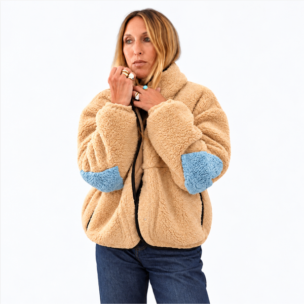 A woman wearing a beige sherpa fleece jacket with light blue elbow patches, paired with blue jeans, photographed against a solid white background.
