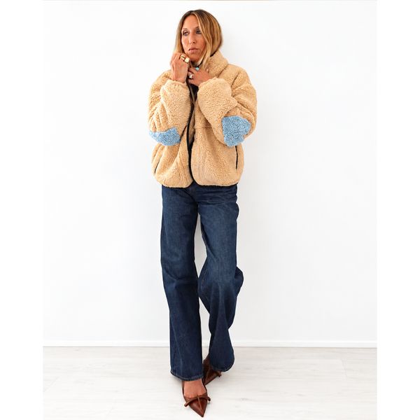A woman stands against a white wall wearing a beige sherpa fleece jacket with light blue elbow patches, dark blue jeans, and brown pointed flats.