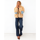 A woman stands against a white wall wearing a beige sherpa fleece jacket with light blue elbow patches, dark blue jeans, and brown pointed flats.